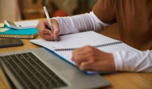 Brazos de persona escribiendo en cuaderno al lado del teclado